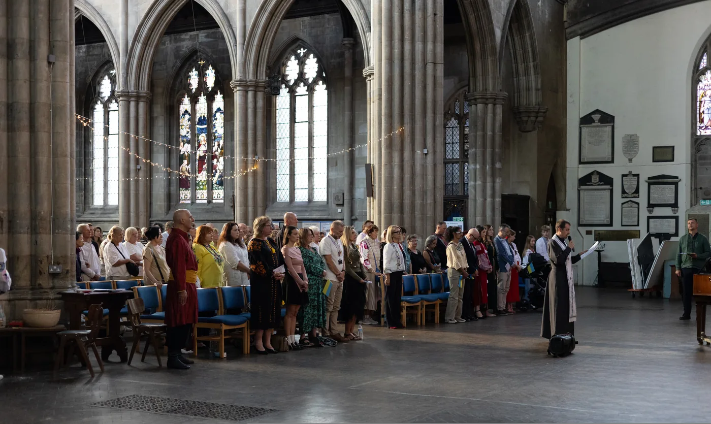 A congregation standing in the nave of a large church with stone pillars, stained glass and strings of lights, facing a robed figure holding a microphone and a sheet of paper
